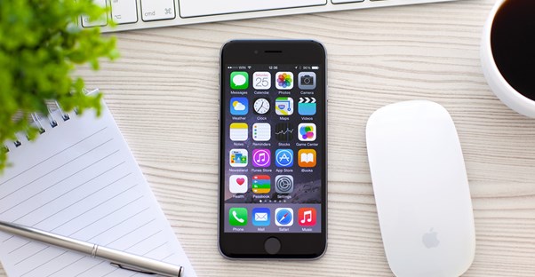 Apple iphone laying on a desk next to a magic mouse and a notebook