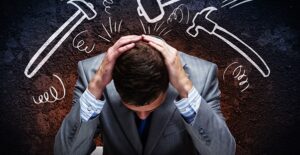 Man in a gray suit sitting at a table with his head in his hands. Chalkboard hammers drawn behind him that seem to be hammering down on him because of the stress of declaring bankruptcy
