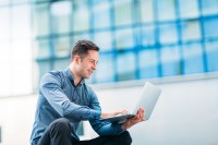 a young man in business casual sitting in front of a nice building with a lot of windows studying on a laptop about successful entrepreneurs