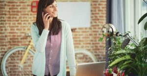 a single mom in business casual standing at her home desk speaking on the phone with a happy look on her face because she got a grant for her small business