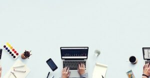 a shot from above of a woman's hands on her laptop keyboard working on application for a business grant with paper and pens and a drink and her phone around the laptop