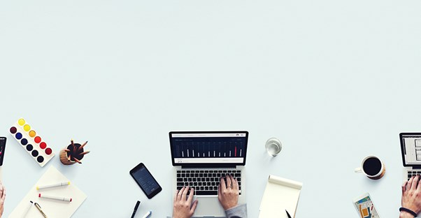 a shot from above of a woman's hands on her laptop keyboard working on application for a business grant with paper and pens and a drink and her phone around the laptop