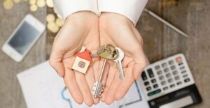 a man's hands cupped and holding two keys with a house shaped keychain on them out over a desk with a home blueprint, a phone, a calculator, a pen, and a pile of cold coins