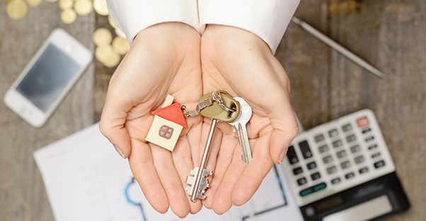 a man's hands cupped and holding two keys with a house shaped keychain on them out over a desk with a home blueprint, a phone, a calculator, a pen, and a pile of cold coins