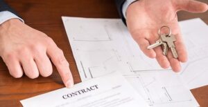 A man with his hands on his desk; one hand points to a white piece of paper with "contract" printed out on it and other hand hovers above a few printed blue prints offering up keys to a home