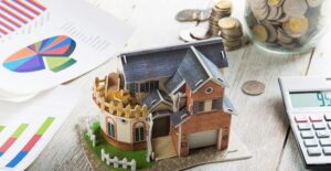 a small play house sitting on a table along with a calculator, a clear jar full of coins, and a piece of paper with a few colored graphs printed on it.