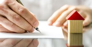 Signing papers next to a mini house. Mortgages for manufactured homes.