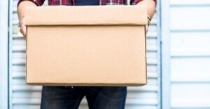 A man holding a cardboard box in front of the metal door of a storage unit