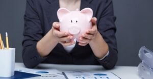a woman behind a desk of financial information holds a piggy bank