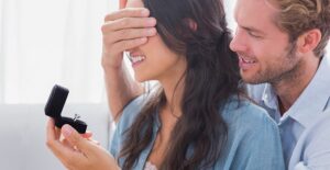 A man covers the eyes of his girlfriend as he pulls out an engagement ring and wonders who will pay for their wedding.