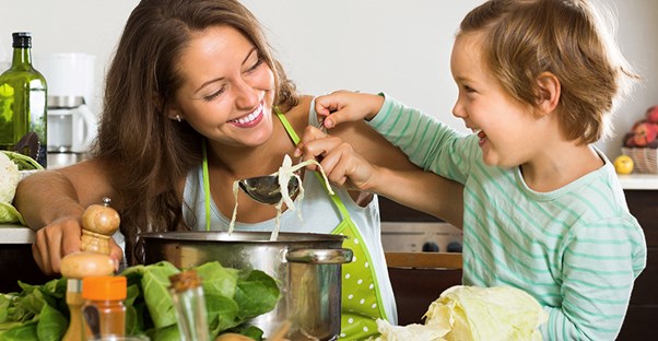 A woman and her child mix ingredients into a large cooking pot surrounded by vegetables, bread, and cheese on the counter.