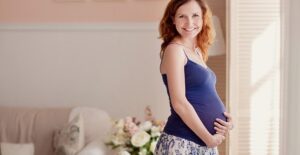 A pregnant woman stands near a sun-lit window in her house holding her stomach.