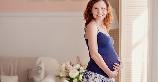A pregnant woman stands near a sun-lit window in her house holding her stomach.