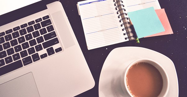 A laptop, a planner, and a cup of coffee sit on a desk