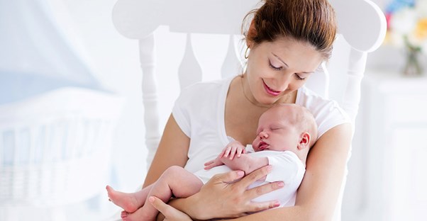 A woman sits in a rocking chair rocking her baby.