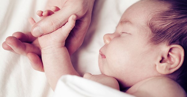 A swaddled baby holds onto the hand of his dad.