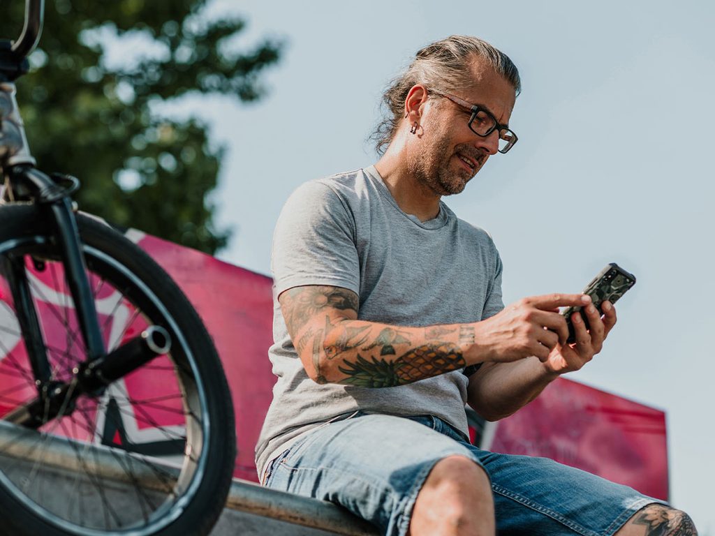 A middle-aged tattooed man looking at his phone while outside at a skatepark.