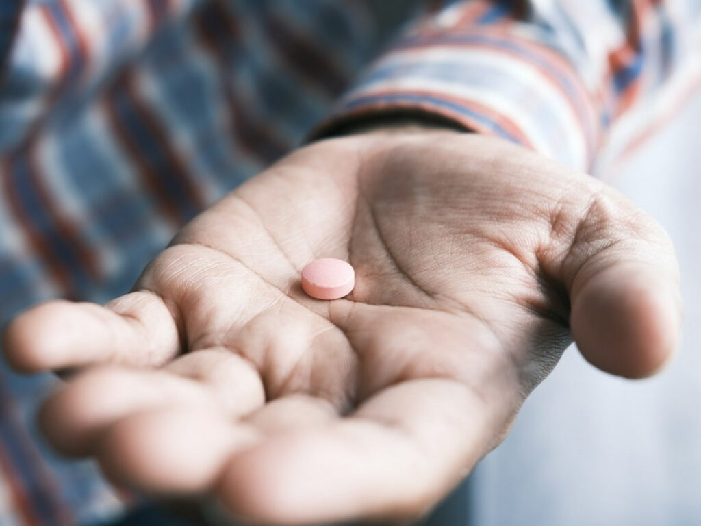 The palm of a man's hand holding out a pink circular pill to the camera.