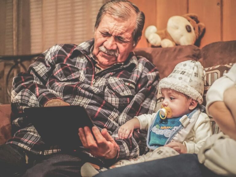 A grandfather in a plaid shirt sitting next to his grandchild baby showing him something on a tablet.