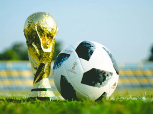 A World Cup soccer ball in front of the World Cup trophy on a field.