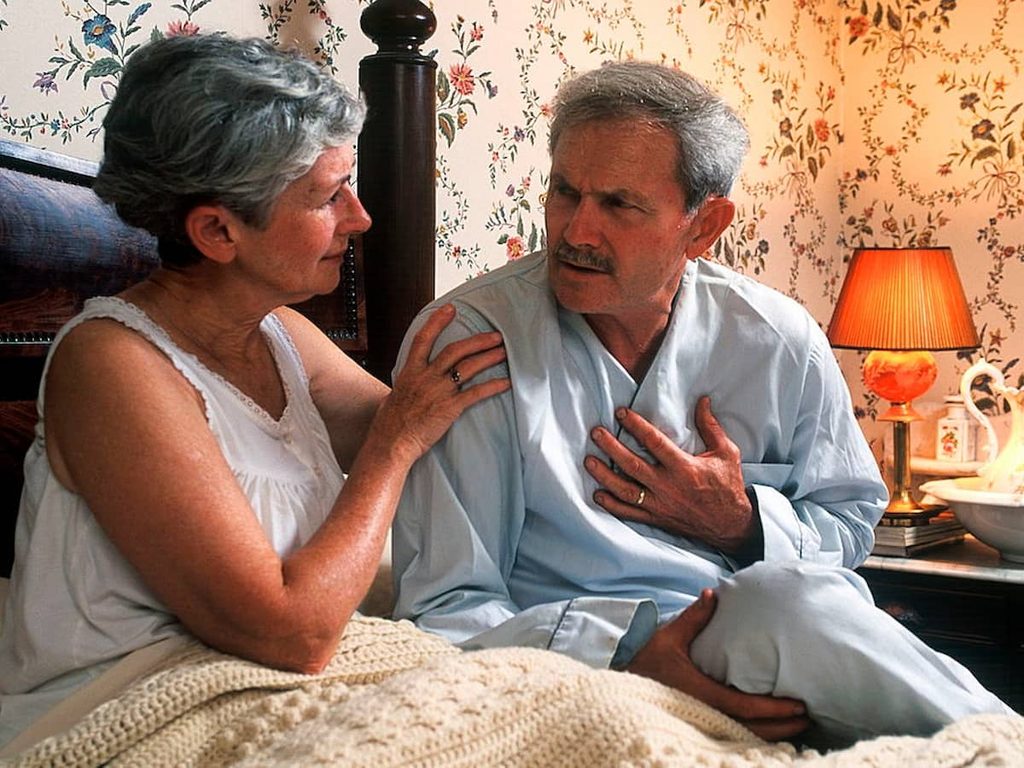 A senior man with chest pain in bed is comforted by his concerned wife.