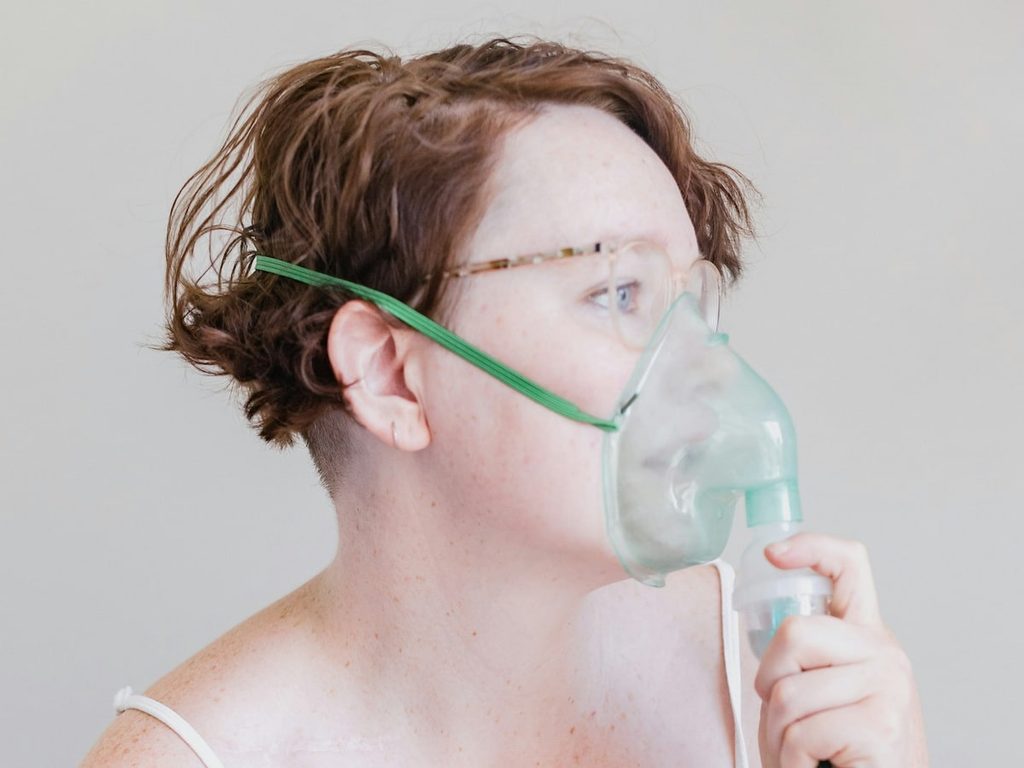 A woman in a white tank top holds an oxygen mask up to her nose and mouth.