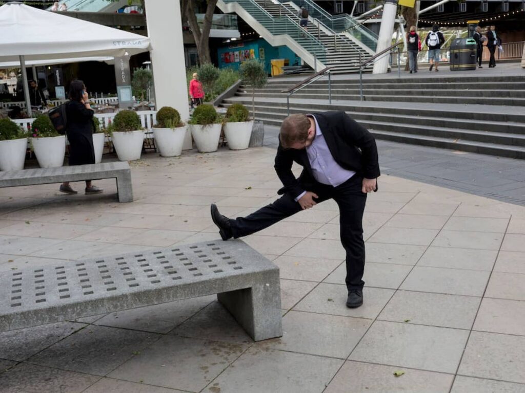 A man wearing a suit stretches the hamstring muscle of one leg in a public plaza.